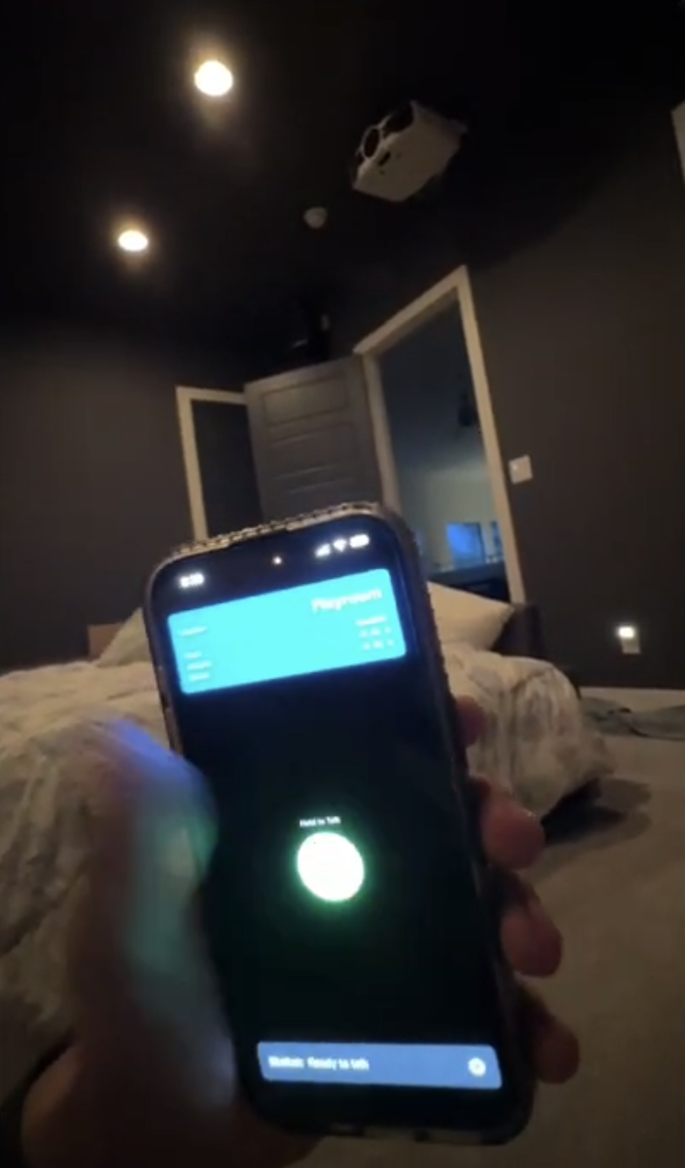 Phone held in a dark bedroom running the Gemi assistant interface while the ceiling lights are on and the room is visible behind it