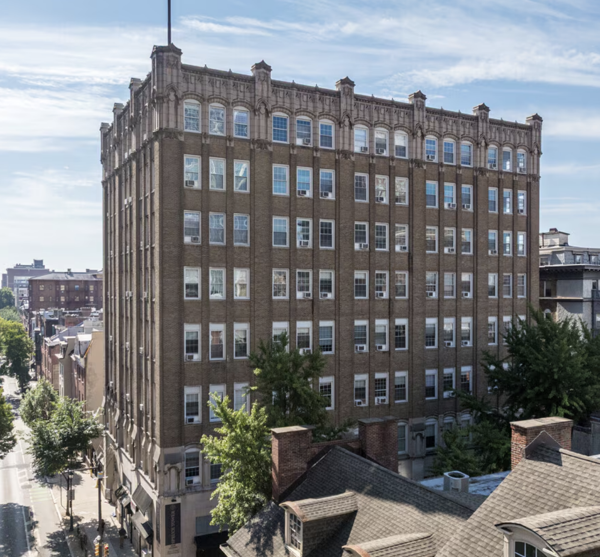 Exterior photo of Spruce Tower — tall historic brown-brick apartment building on a Center City Philadelphia corner, with ornate cornice details, rows of white-trim windows, and blue sky overhead