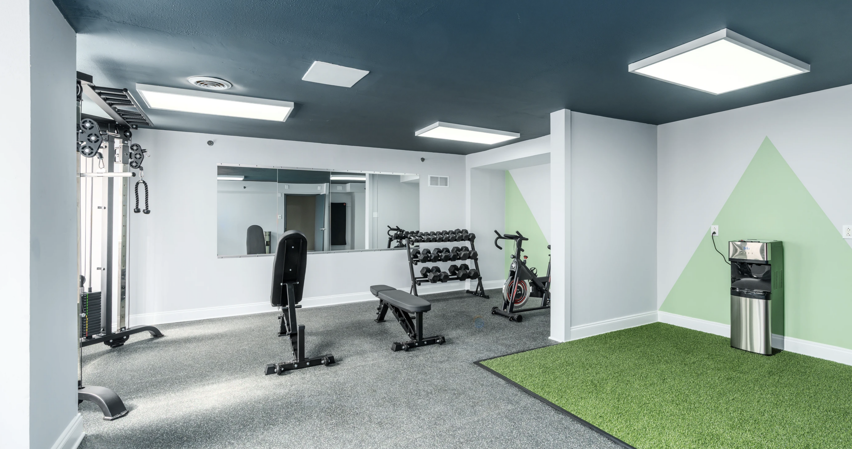 Final real amenity photo of the Spruce Tower gym — bright white room with mirrored wall, cable machine, weight bench, dumbbell rack, exercise bike, green turf, and pale green wall graphics