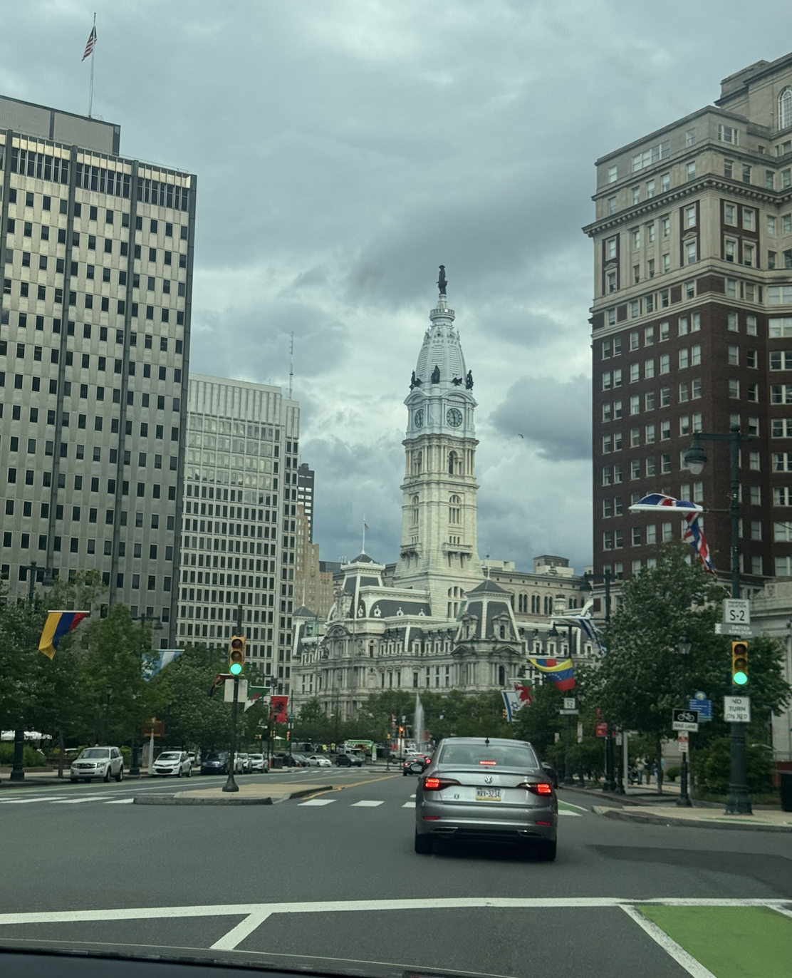 Photo through the windshield approaching Philadelphia City Hall — Broad Street traffic leading toward the clock tower under heavy clouds, framed by tall Center City buildings